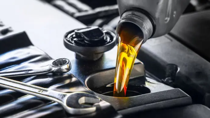 A close-up shot showing fresh, golden-amber engine oil being poured from a silver plastic container into a car engine's oil fill port. Two silver wrenches rest on the black engine cover in the foreground, next to the open oil cap.