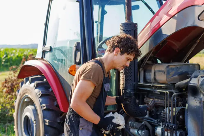 A man with curly brown hair wearing a brown t-shirt and blue overalls is leaning over the exposed engine of a large red tractor in a field, appearing to work on it or clean it with a white cloth.