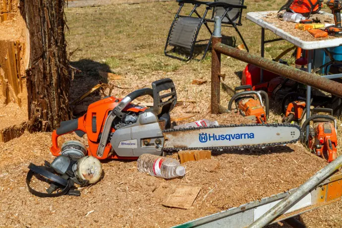 An orange and white Stihl chainsaw sits resting on a pile of fresh wood chips and sawdust. The saw's bar, which features the Stihl logo, points toward the right side of the frame. In the background, a person wearing an orange shirt is partially visible, and more logs are stacked behind the equipment.