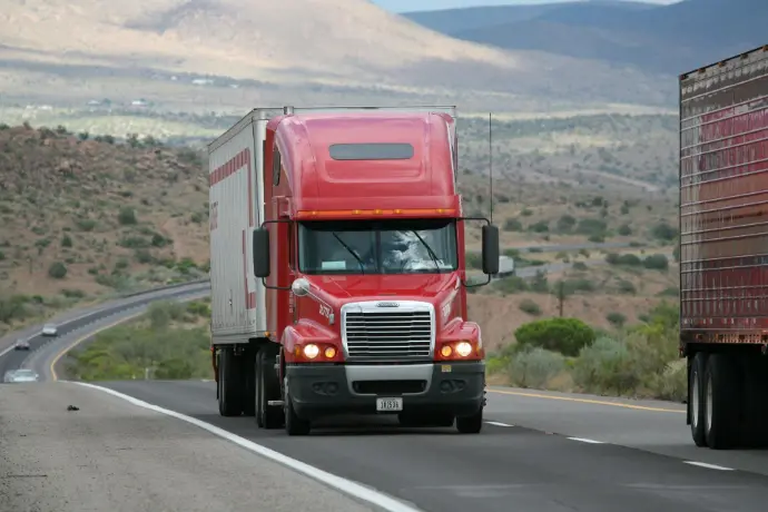 A red semi-trailer truck travels down an open highway through a desert landscape. The bright red cab of the truck stands out against the muted tones of the road and the distant hills. The trailer behind it is a neutral white or silver, and the vast, open space suggests a long-haul journey.