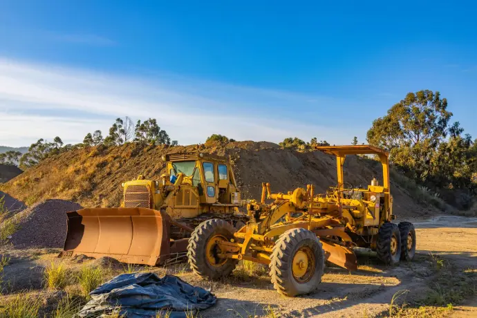 An outdoor shot shows two pieces of heavy yellow machinery parked in a construction or mining site. On the left is a bulldozer with a large metal blade and a cabin, and on the right is a motor grader featuring six large tires and a long frame. They sit on a dirt ground with a large pile of earth and some trees in the background under a clear blue sky.