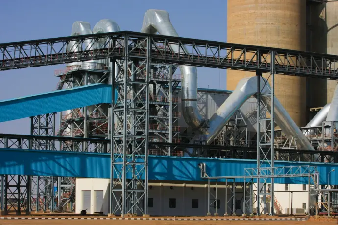 An industrial plant with large silver pipes, steel scaffolding, and a tall tan tower. Blue covered conveyors stretch horizontally across the foreground and midground under a clear sky.